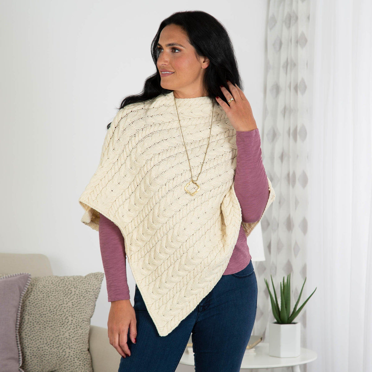 Woman wearing a cream-colored knitted poncho in a home setting