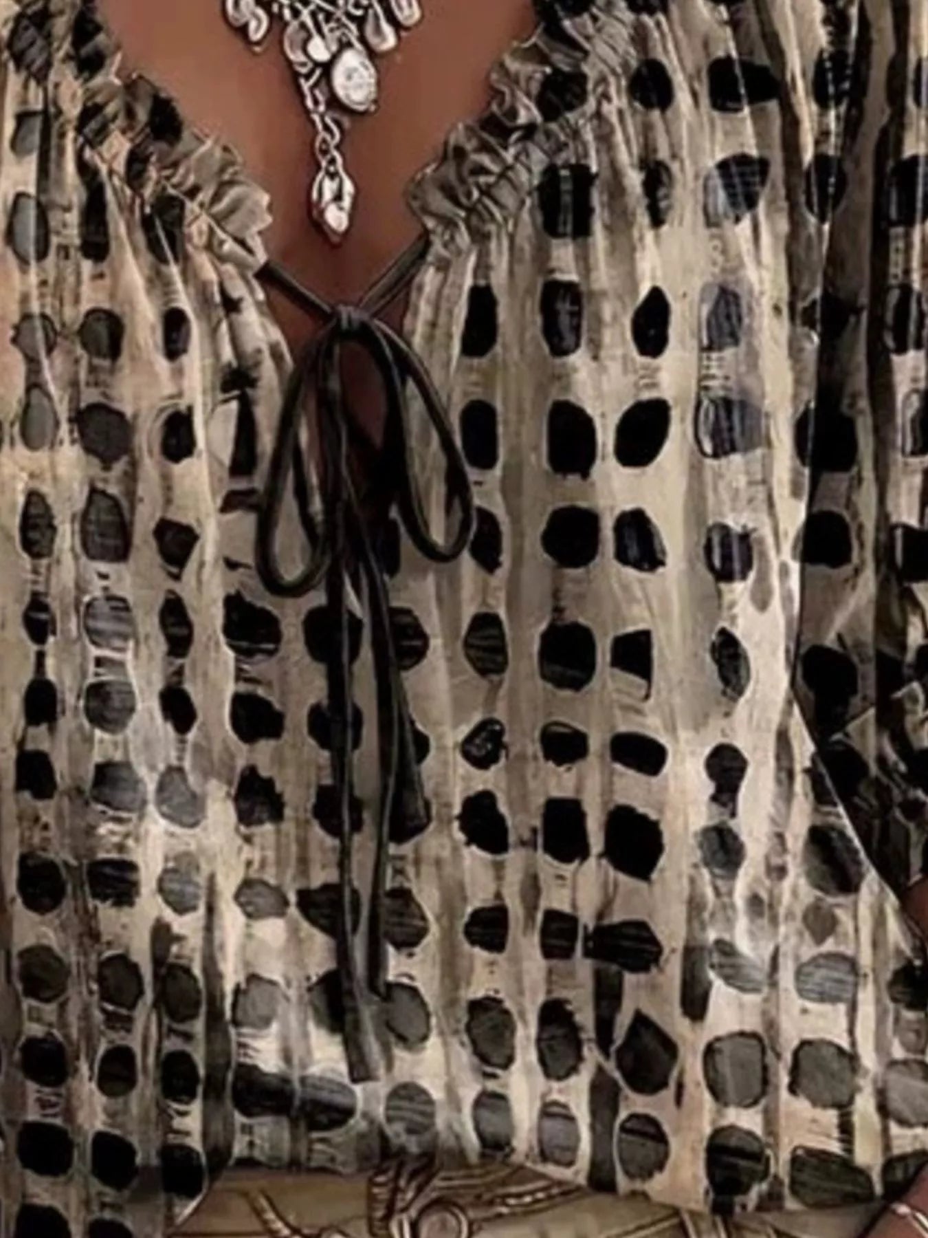 Close-up of a women's blouse with a beige and black abstract spotted pattern, featuring a ruffled V-neckline and a black tie closure at the center chest.