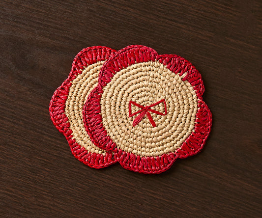 Handwoven raffia fringe coasters in red and natural colors with a bow design, set on a dark wooden table.