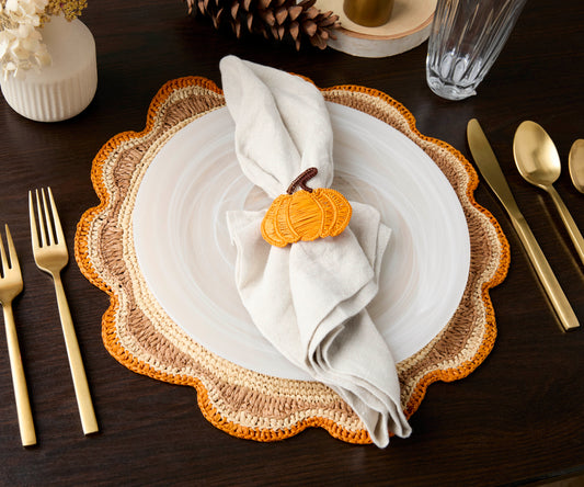 Round raffia woven placemat in crochet style with orange and natural tones, set on a dark wooden table with gold utensils.