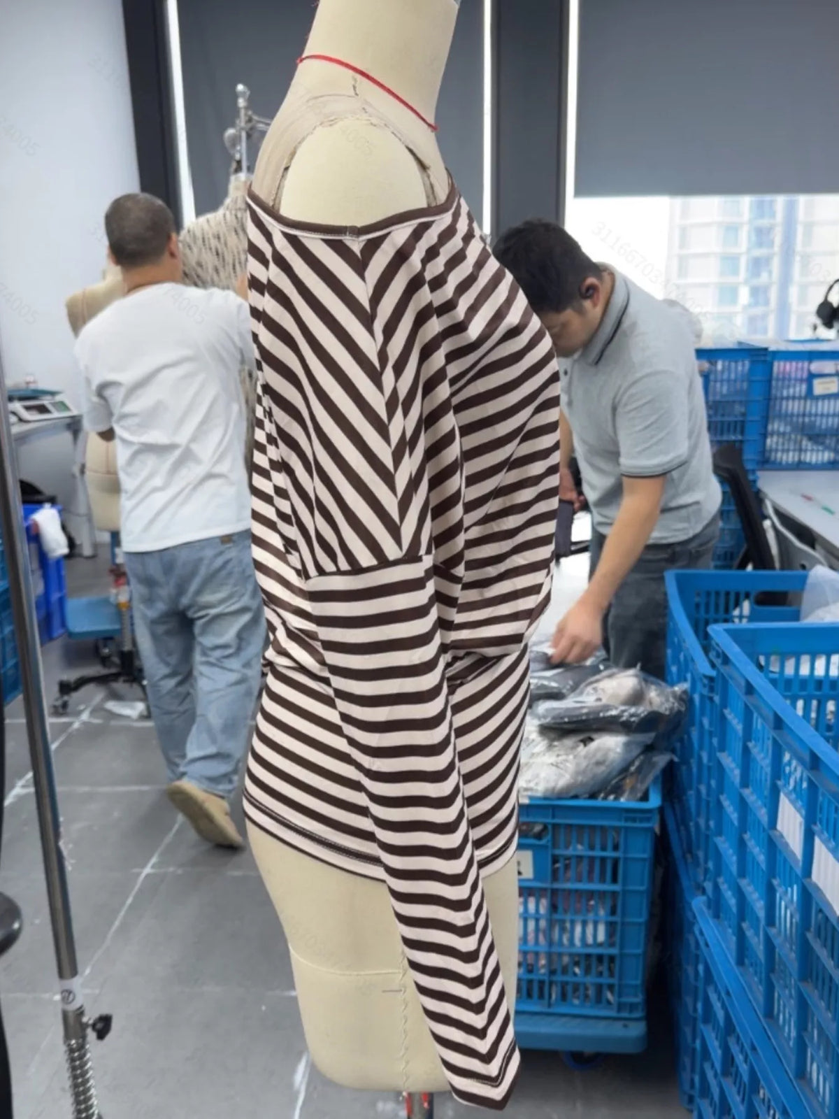 A brown and beige horizontally striped knit top displayed on a dress form mannequin in a warehouse or tailoring workspace, with two men working in the background near blue crates.