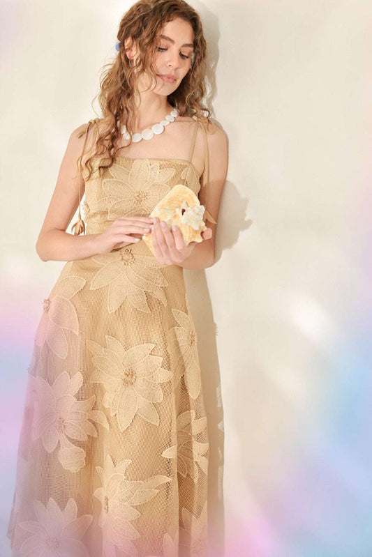 A woman wearing a champagne beige floral tulle dress and holding a matching beige clutch bag, set against a soft, light-colored background with pastel light leaks.