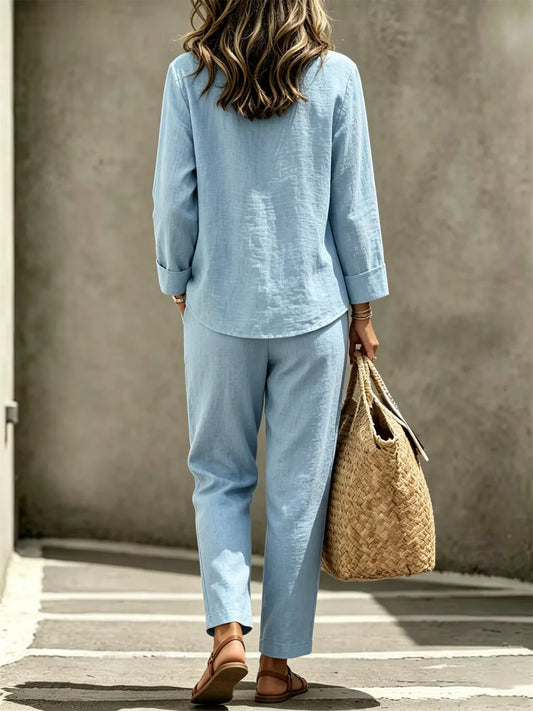 A woman viewed from behind wearing a light blue linen two-piece set consisting of a long-sleeve button-up shirt and matching relaxed-fit pants. She is holding a large woven straw tote bag and wearing brown leather sandals, standing against a textured gray concrete wall.