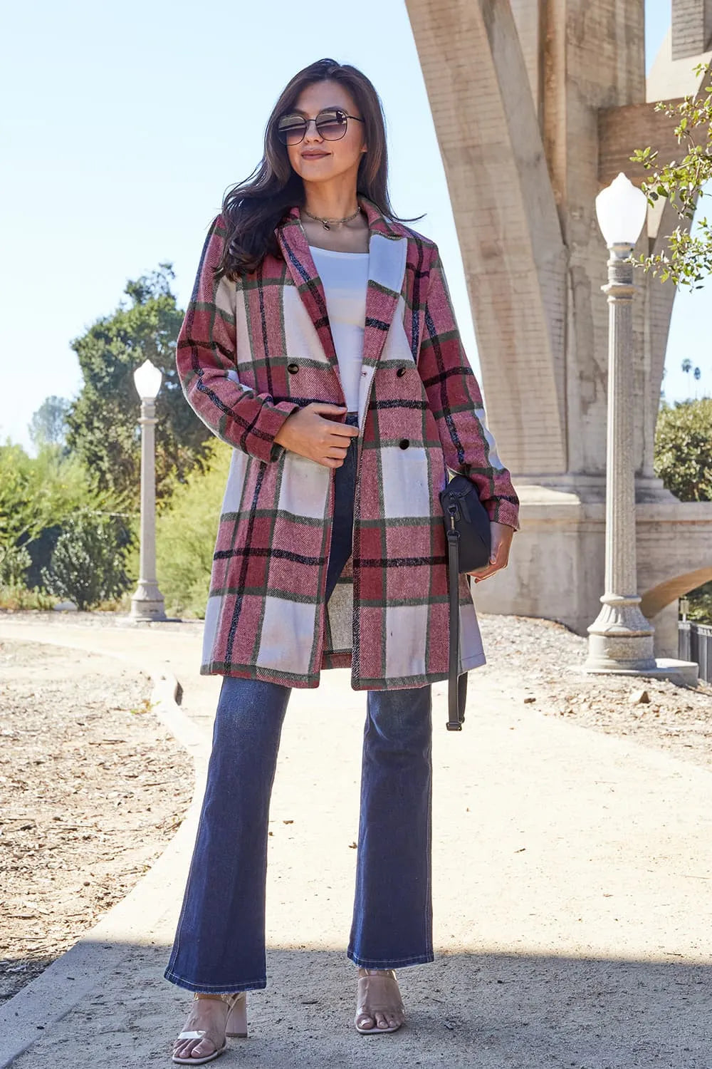 Woman wearing a plaid coat and blue jeans standing outdoors with architectural elements in the background.