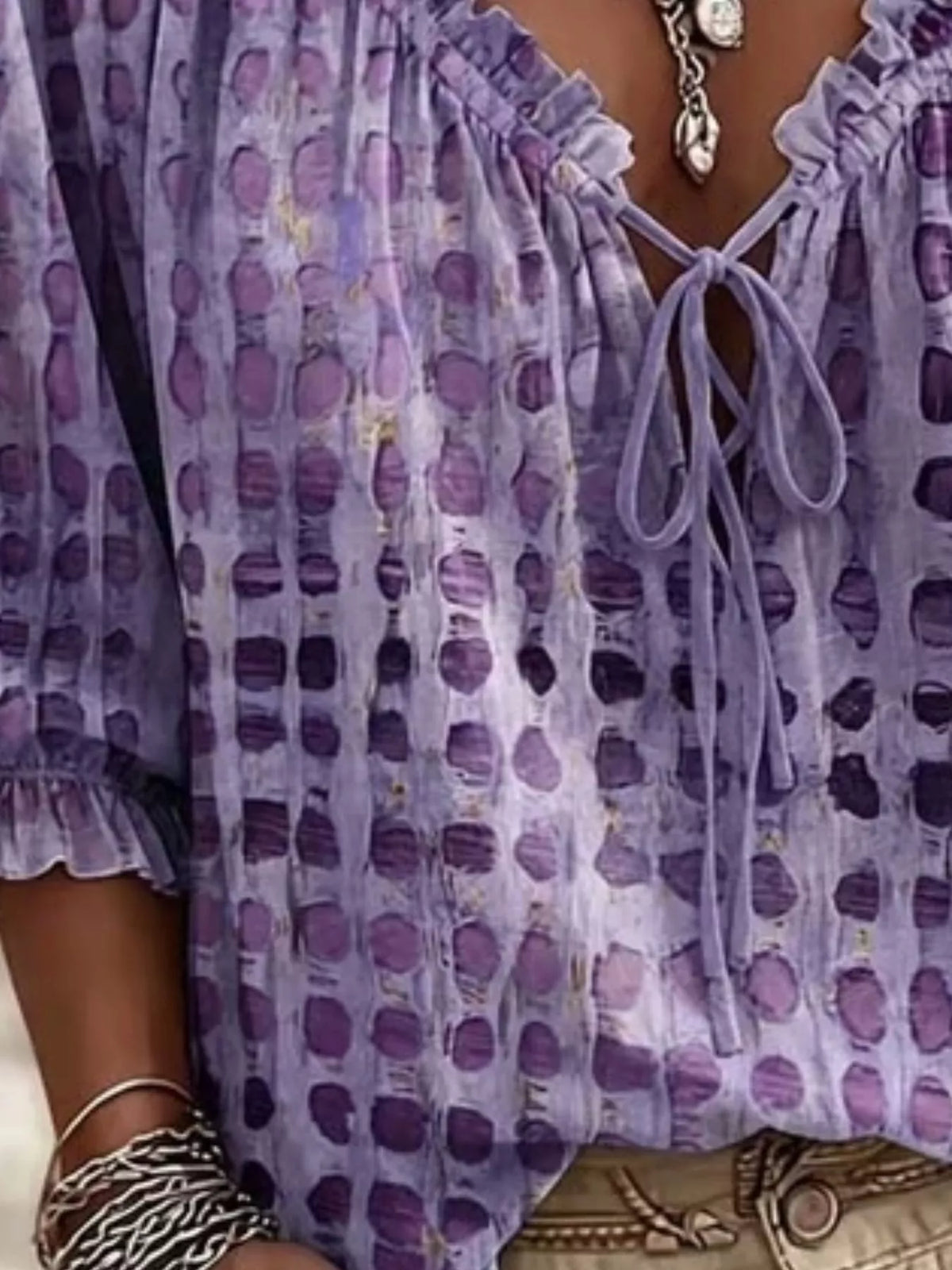 A close-up of a person wearing a lavender and purple patterned blouse with a ruffled neckline, tie-front closure, and elbow-length sleeves. The blouse has an abstract dotted or watercolor-like print in shades of purple and lavender.