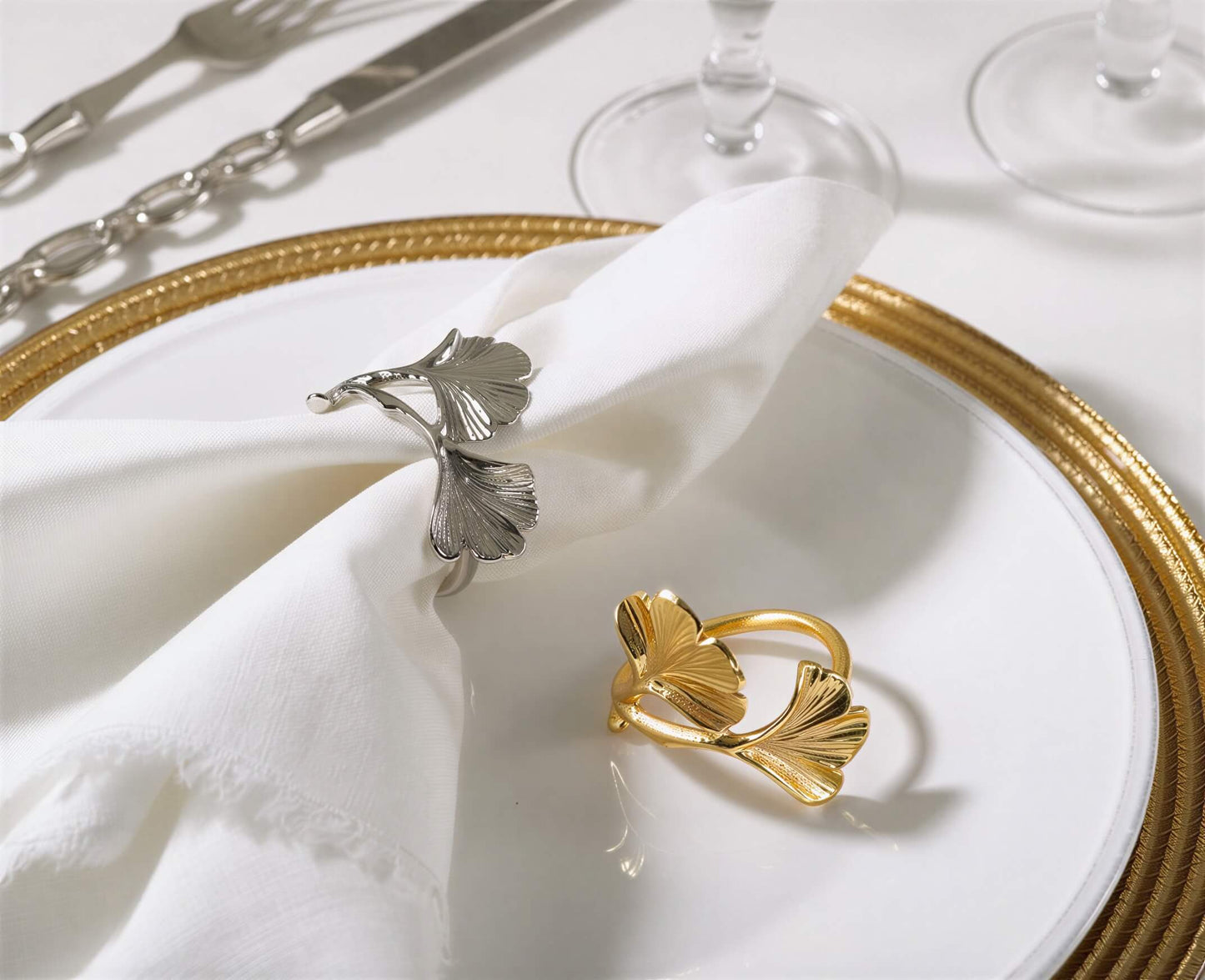 One silver and one gold metal ginkgo leaf napkin ring on a white ceramic plate with a white fabric napkin.