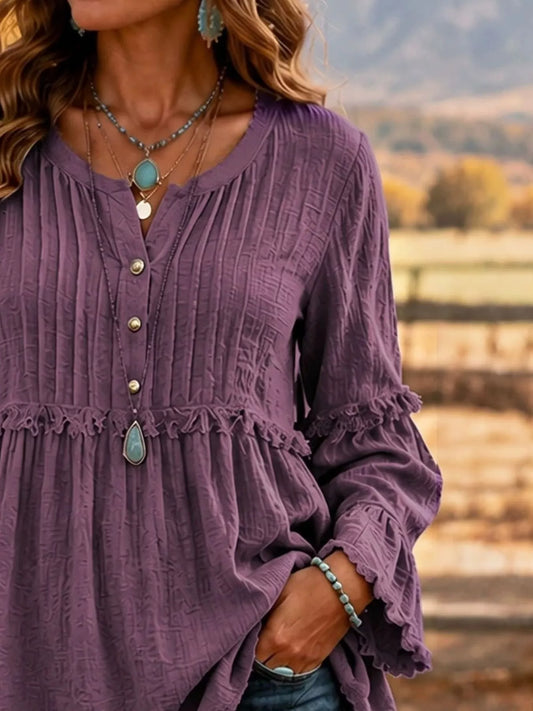 A woman wearing a textured purple long-sleeve blouse with a buttoned yoke and ruffled details, photographed outdoors against a blurred natural background.