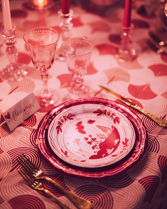 Stacked red and white paper plates with floral and pavilion patterns on a patterned tablecloth with glassware and cutlery.
