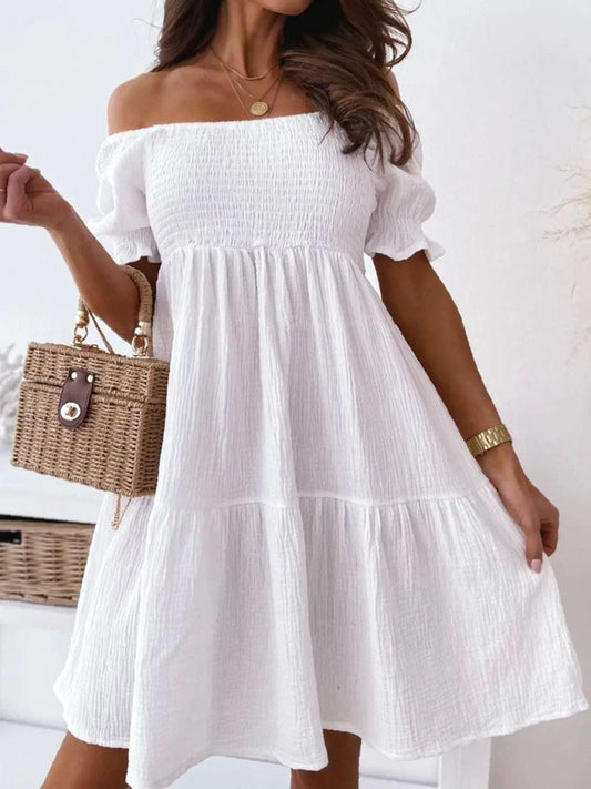 A woman wearing an beige off-shoulder mini dress with smocked detailing, paired with a straw bag and gold accessories.