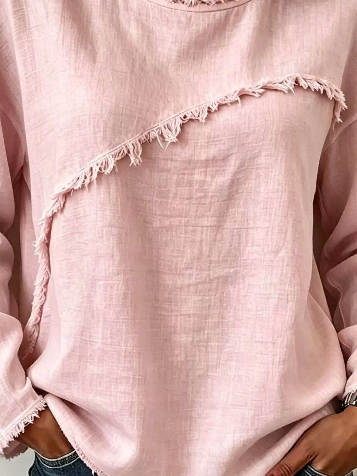 A close-up of a light pink long-sleeve top with diagonal frayed fringe trim, worn by a model.