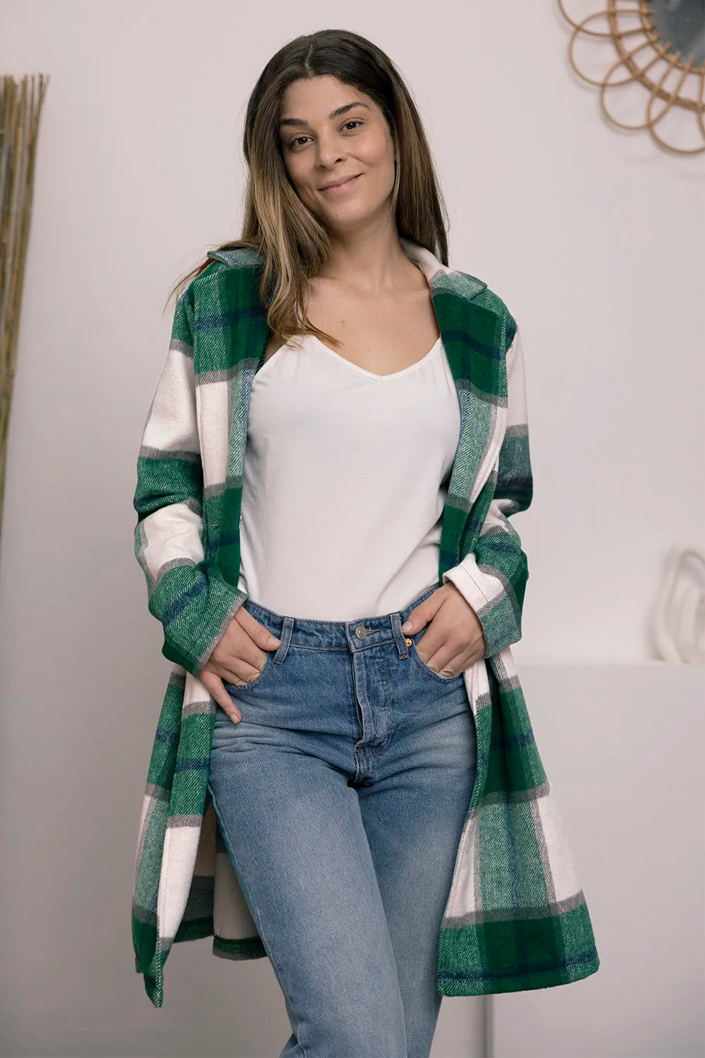 Woman wearing a green and white plaid shirt over a white top and blue jeans, standing against a plain background.