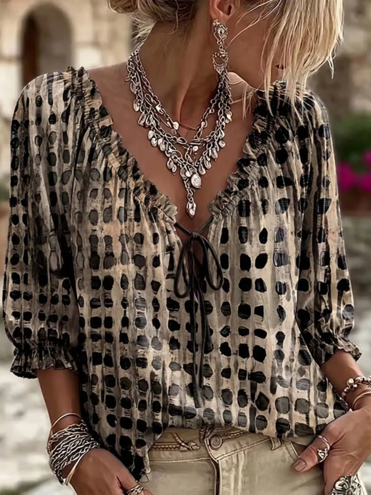 A woman wearing a beige and black leopard-print blouse with a ruffled V-neckline and tie-front detail, styled with layered silver statement necklaces and bracelets.