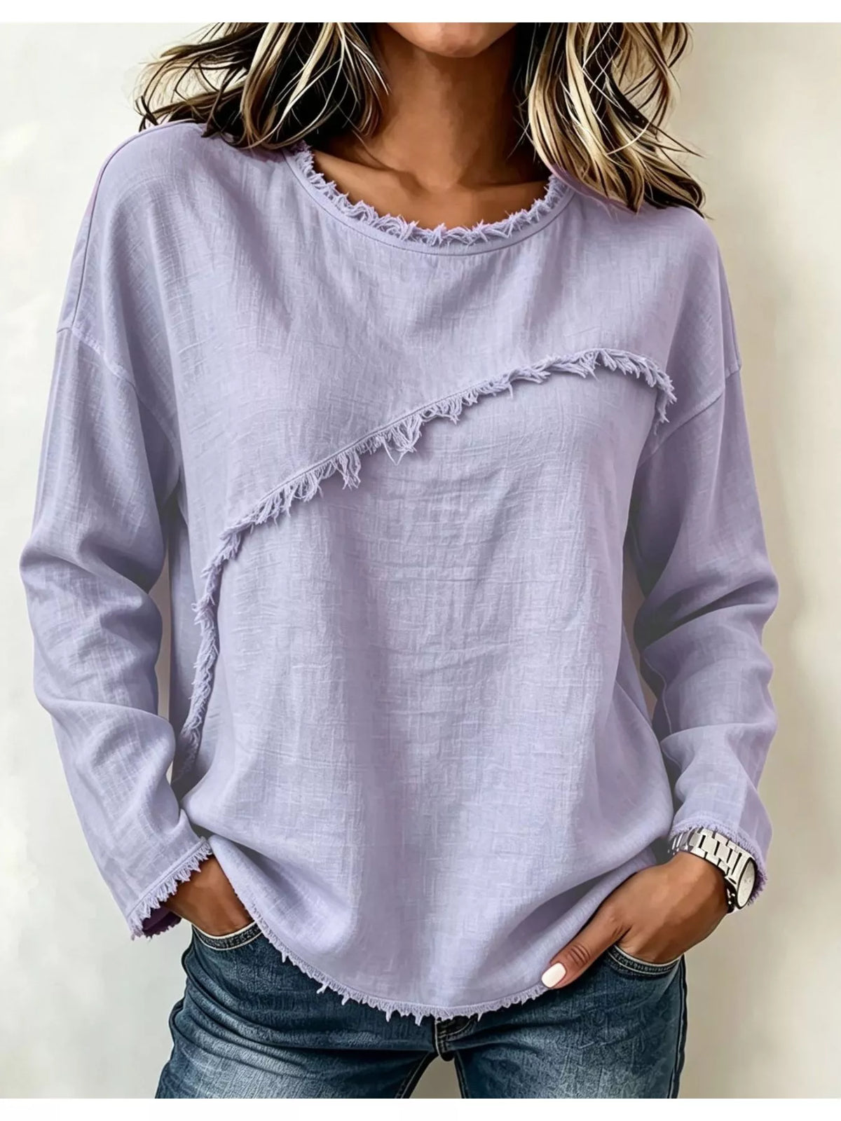 A woman wearing a light lavender long-sleeve top with frayed, raw-edge detailing along the neckline and across the front, paired with blue jeans.