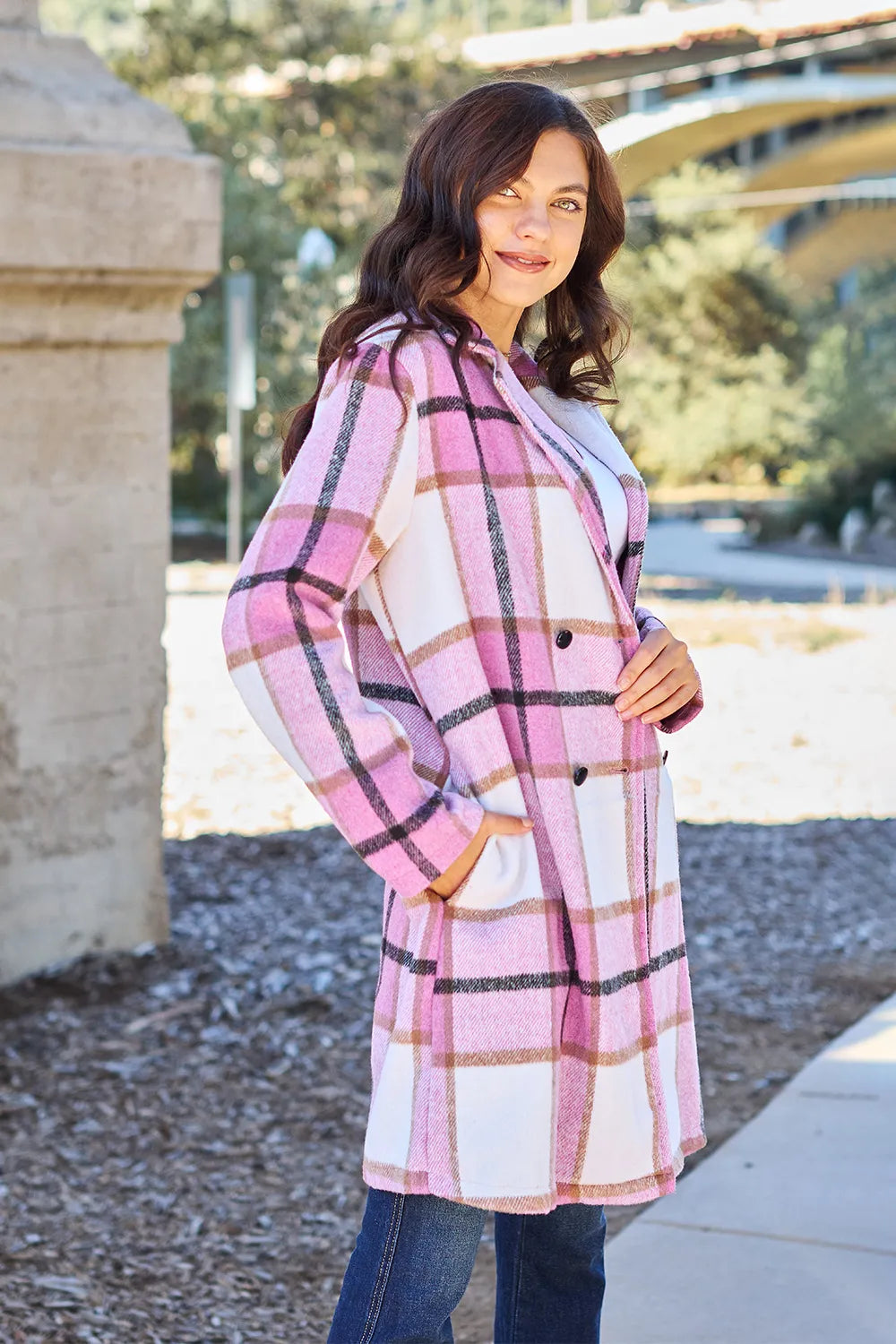 Woman wearing a pink plaid coat standing outdoors with trees and a building in the background.