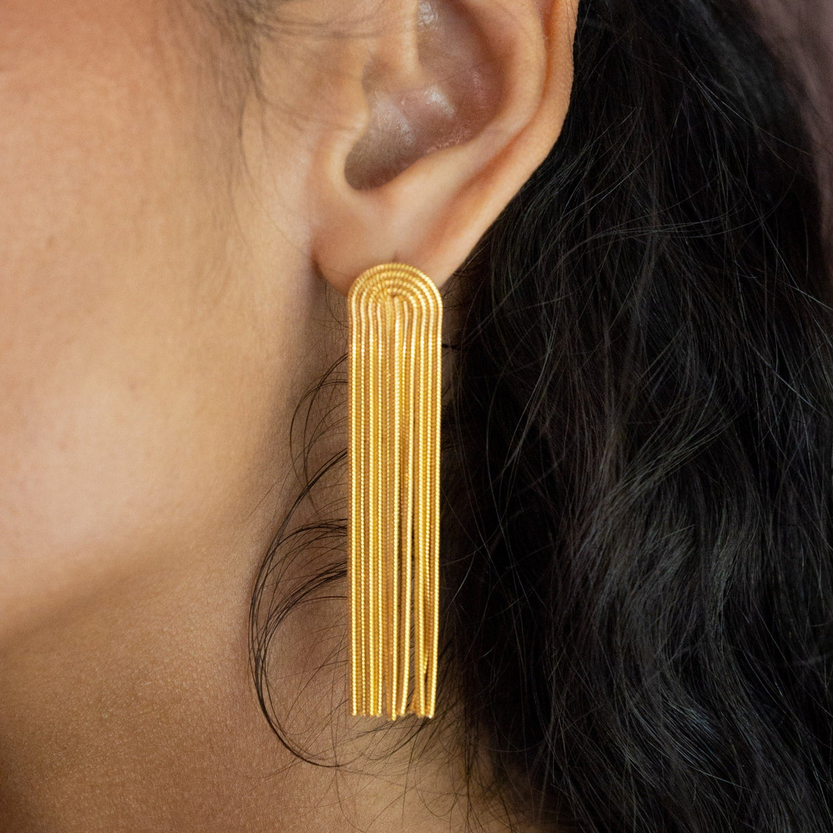 Addison gold statement earring worn with dark hair swept back, showing the long chain fringe beside the jawline.