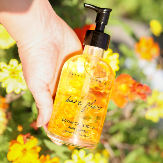 Hand holding a glass bottle of Bare Flower Botanical Body Oil with a black pump against a background of orange flowers.
