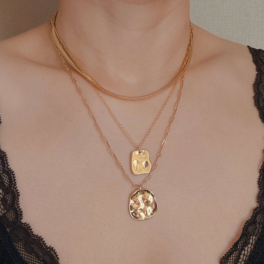Three layered gold metal necklaces including a snake chain, a rectangular pendant, and a hammered round coin pendant.