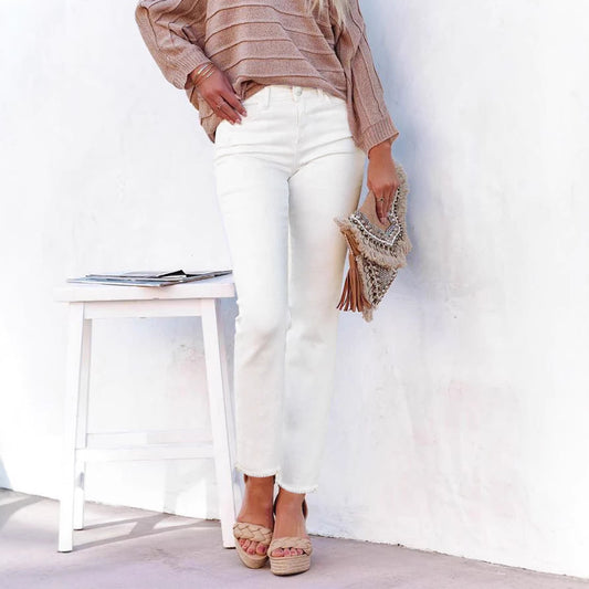 White straight leg denim jeans with a raw hem worn with a tan sweater and wedge sandals against a white wall.
