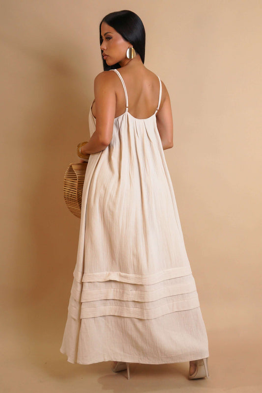 A woman wears a beige fabric maxi dress with thin straps and tiered hem detail, holding a round wooden bag.