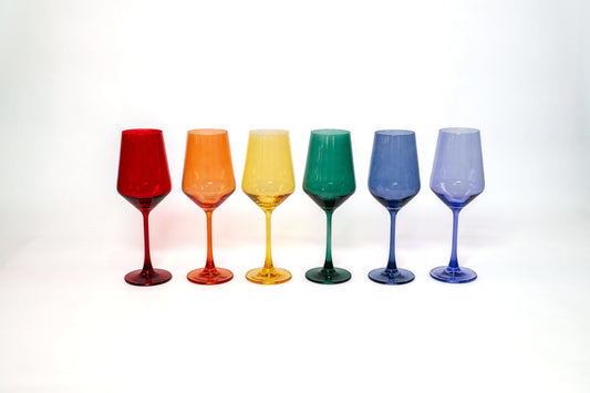 Six glass wine glasses in a rainbow row of red, orange, yellow, green, light blue, and purple on a white background.