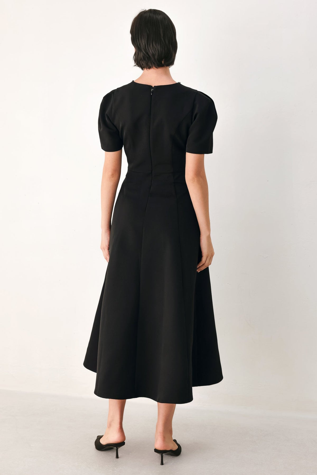 Back view of the black midi dress showing its zipper closure, short puff sleeves and flared midi hem.
