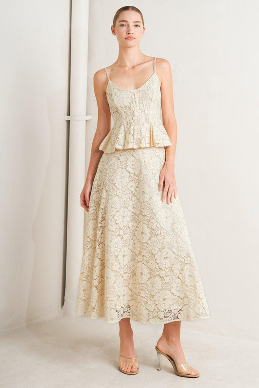 A model wears a matching cream lace camisole top and midi skirt set, styled with clear high-heeled sandals against a minimalist studio backdrop.
