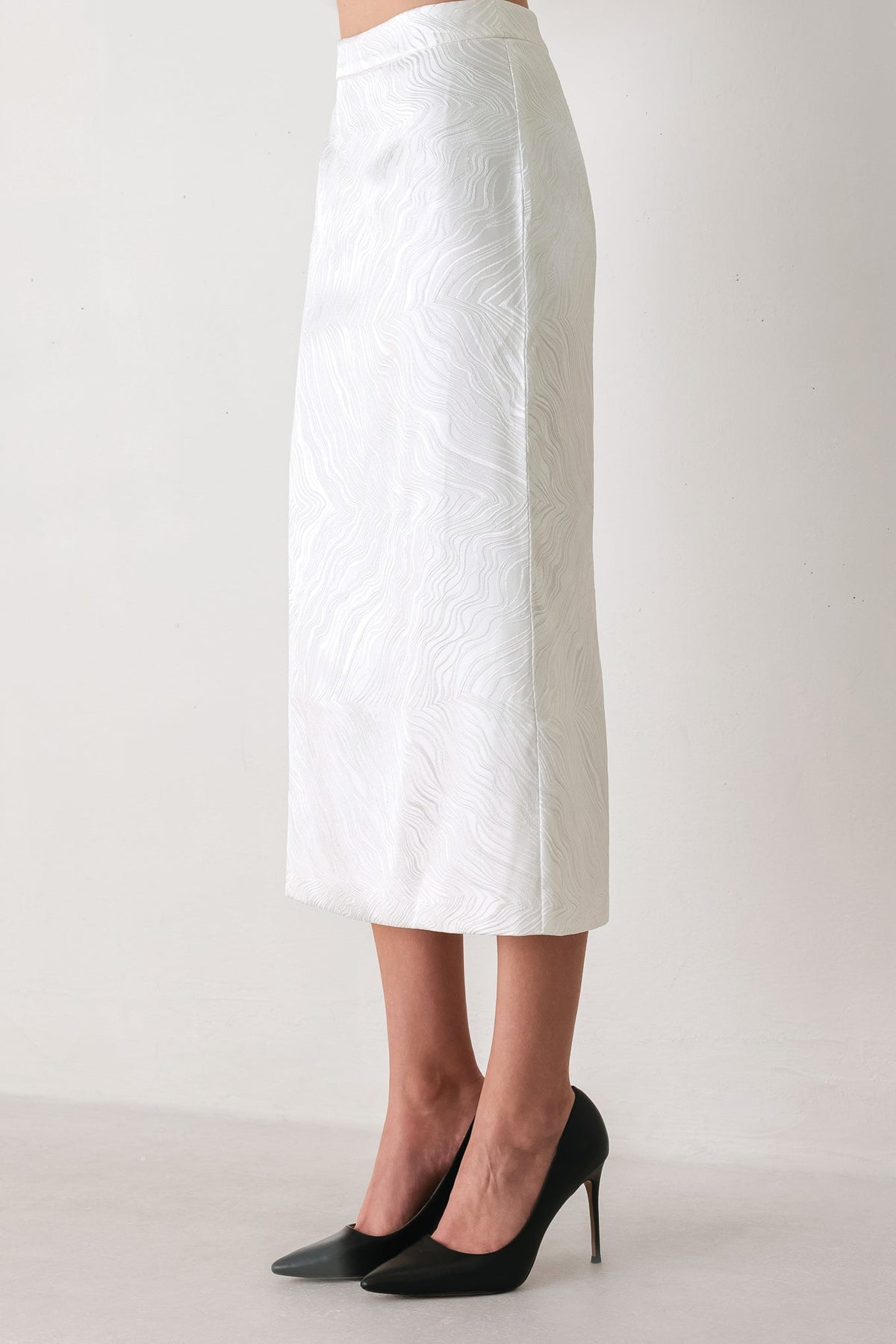 A white midi skirt with a subtle textured leaf pattern, shown on a model wearing black pointed-toe high heels against a plain light background.