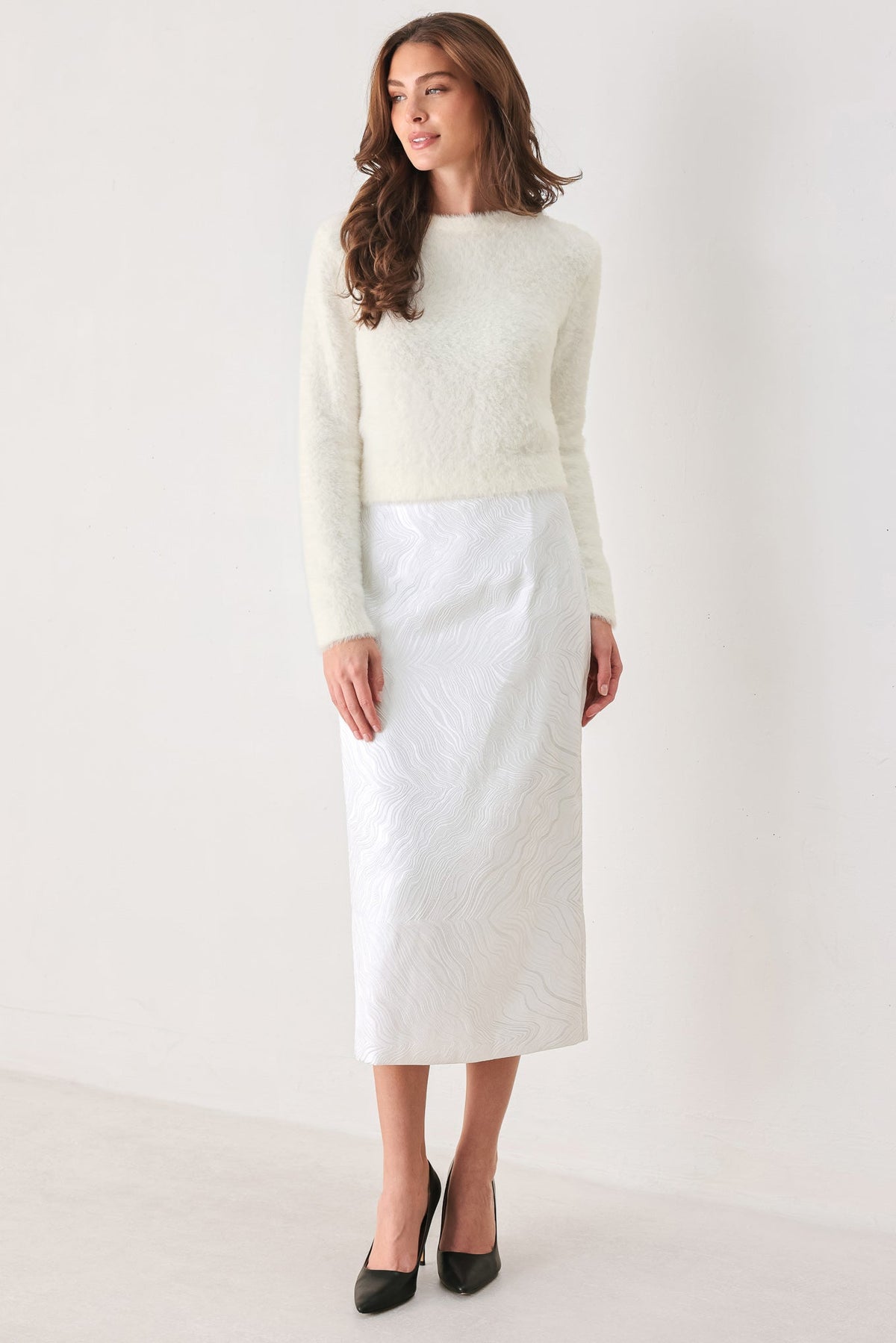 A woman modeling a cream-colored textured knit sweater paired with a white textured midi skirt and black pointed-toe heels, set against a plain off-white studio background.