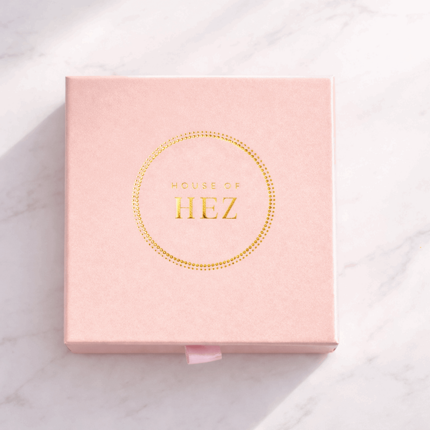 Square pink box with a gold foil HOUSE OF HEZ logo inside a dotted circle, resting on a white marble surface.