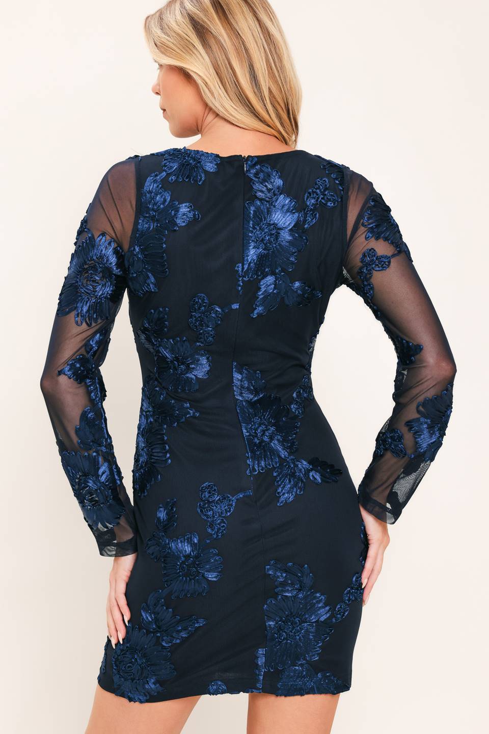A woman shown from the back wearing a fitted mini dress with long sheer mesh sleeves and dark blue floral lace applique over a black base, against a plain light beige studio background.