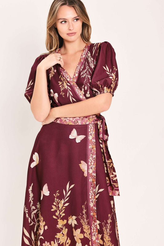 A woman wearing a deep burgundy wrap dress with a gold and white butterfly and floral print, set against a plain light beige background.