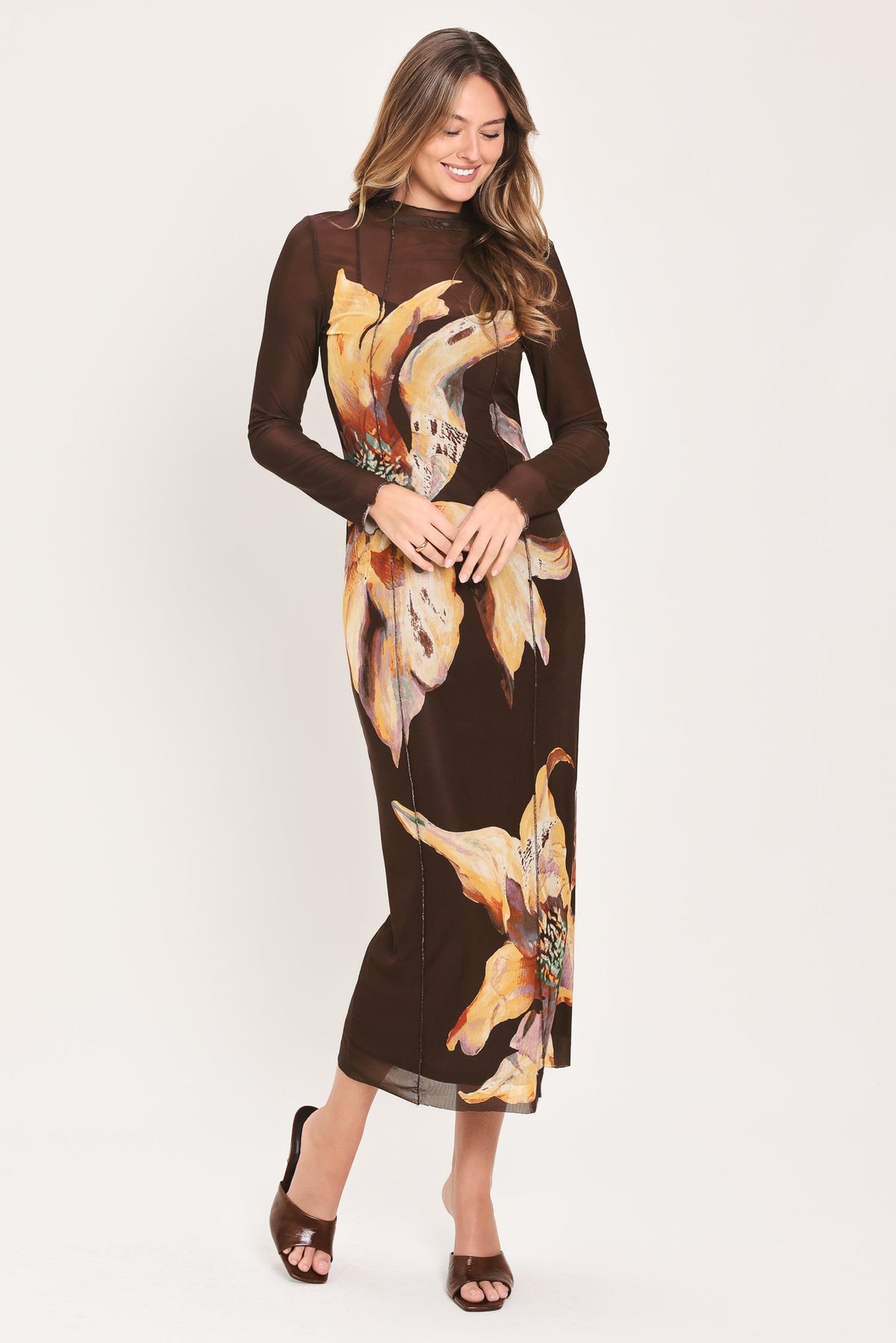 A woman modeling a long-sleeve brown midi dress with a large yellow and orange floral print, paired with brown heeled sandals, against a plain light background.
