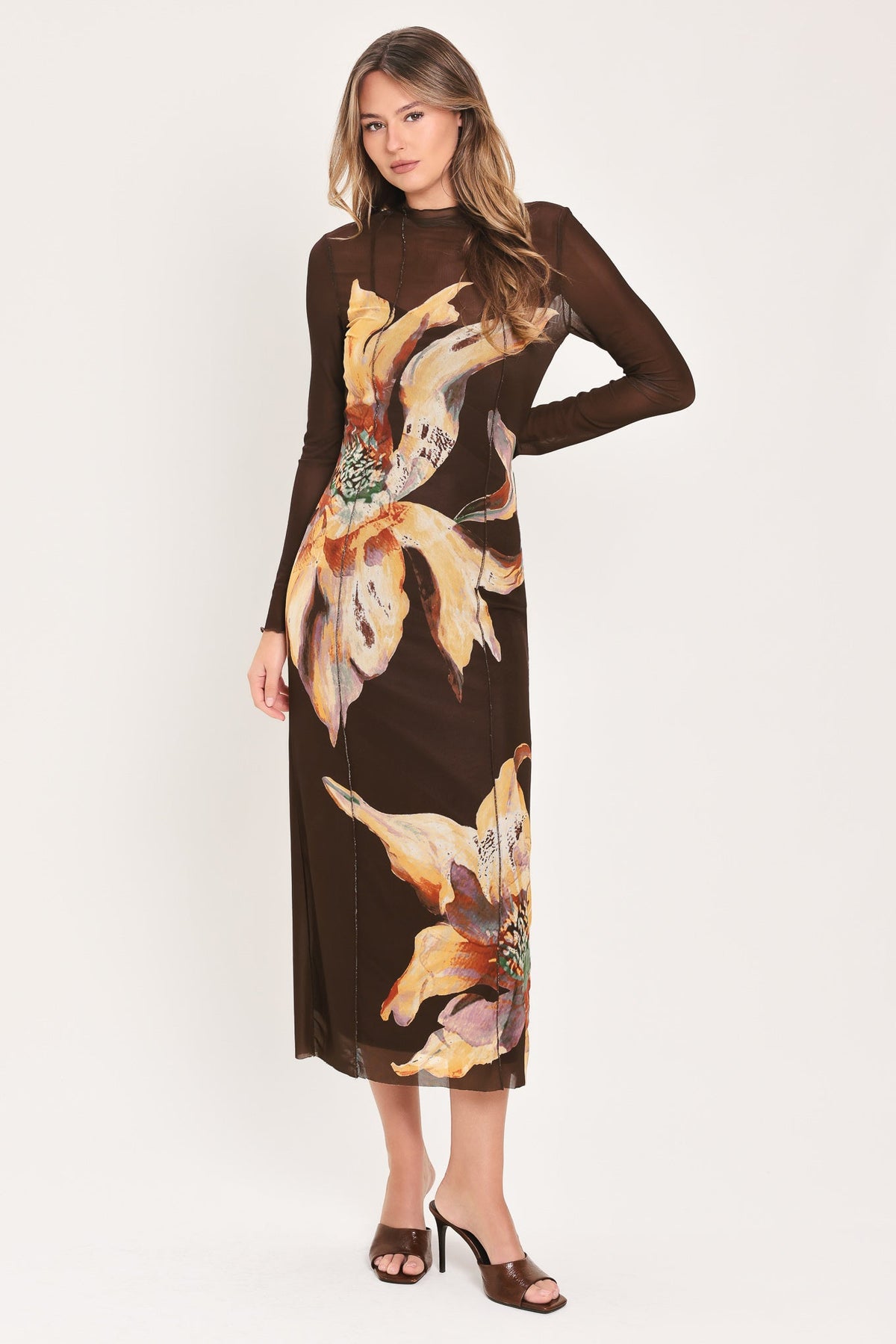 A woman modeling a long-sleeve mock-neck midi dress in dark brown with a large yellow and orange floral print, paired with brown heeled sandals, against a plain white studio background.
