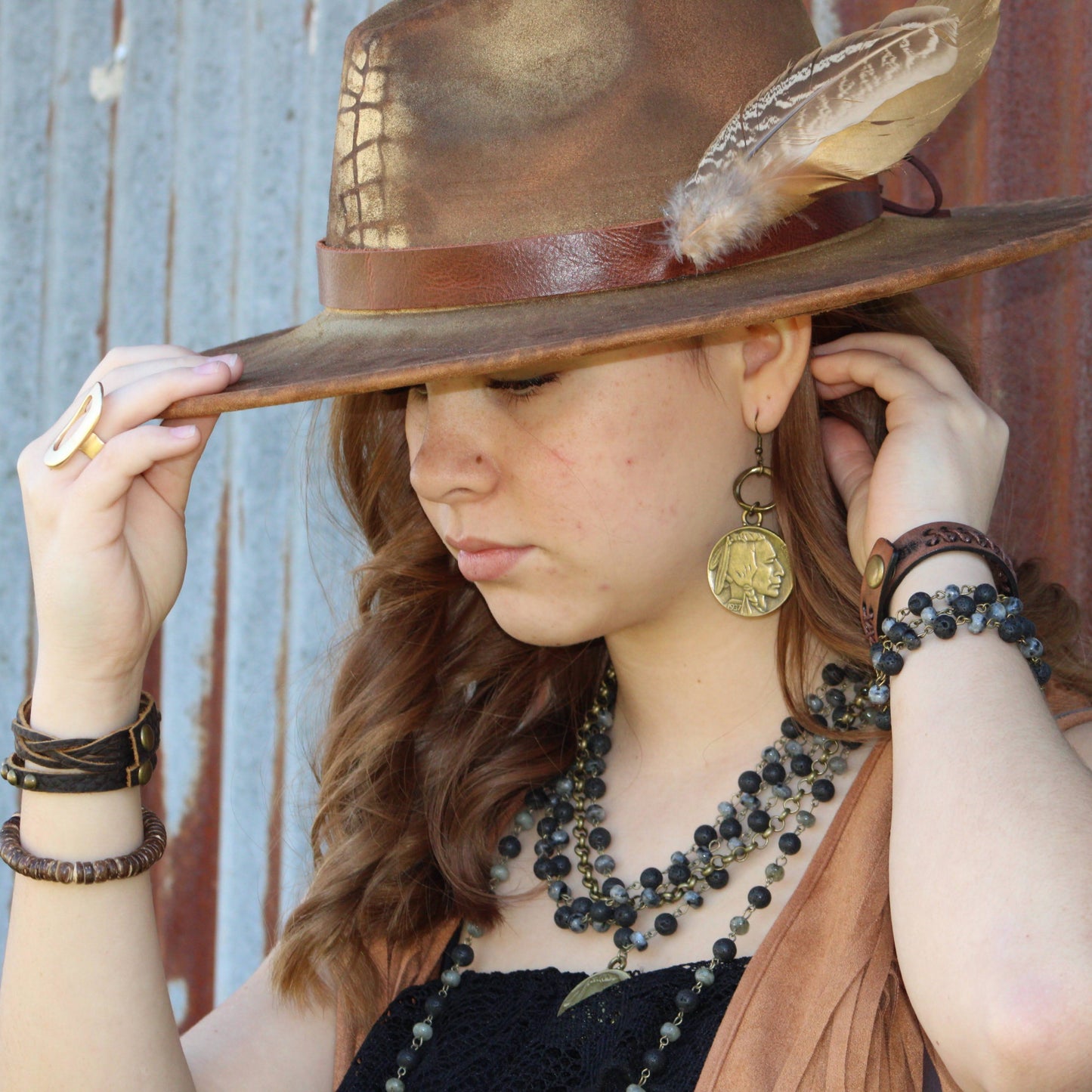 Woman wearing a wide-brimmed hat with a feather, multiple necklaces, and bracelets against a rustic wooden background.