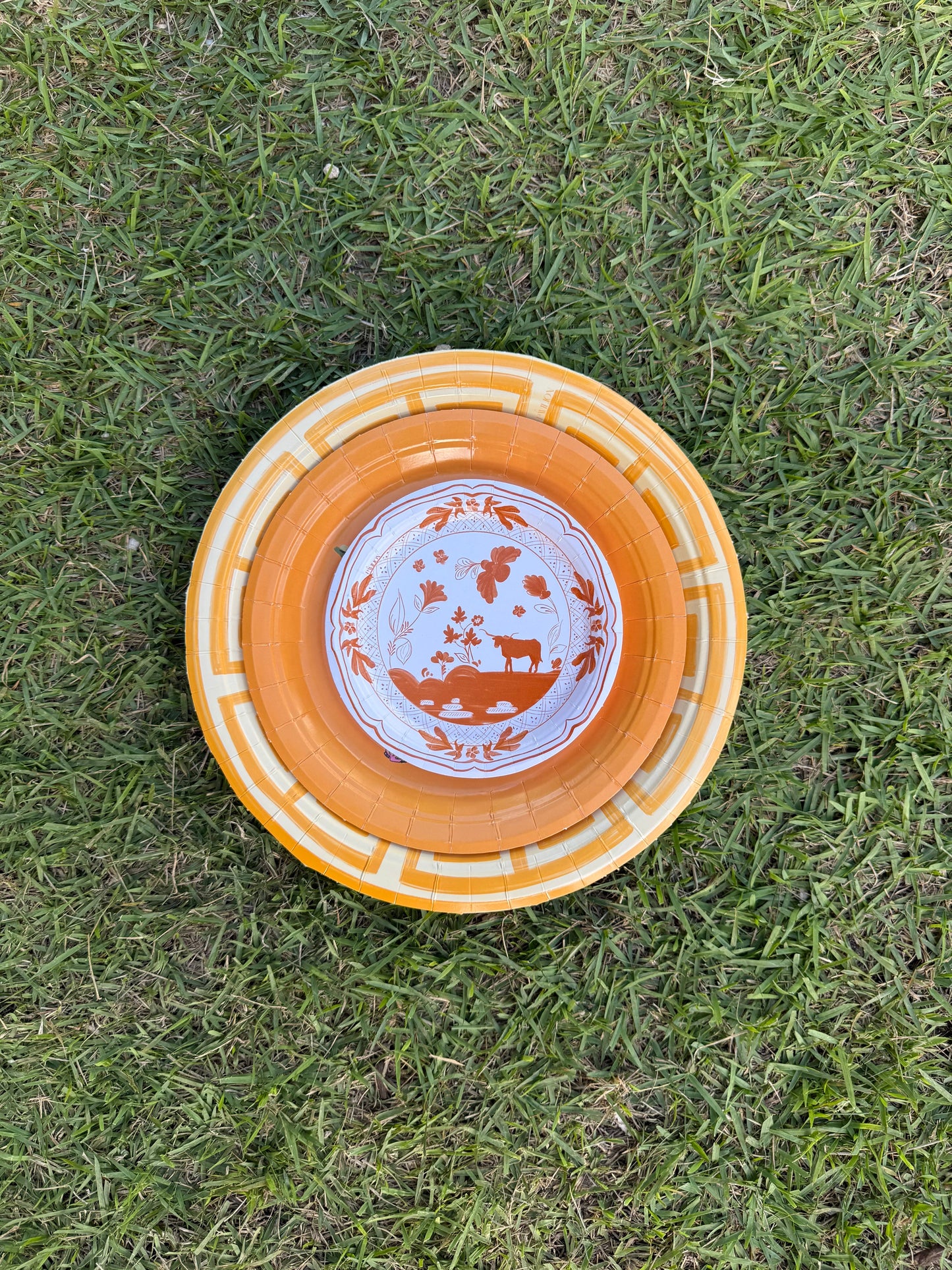 Three stacked yellow and orange paper plates with Greek key and cow motifs on green grass.