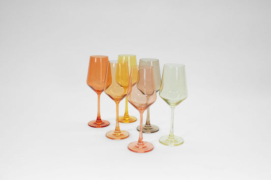 Six assorted orange, yellow, and grey tinted glass wine glasses with stems arranged on a white background.