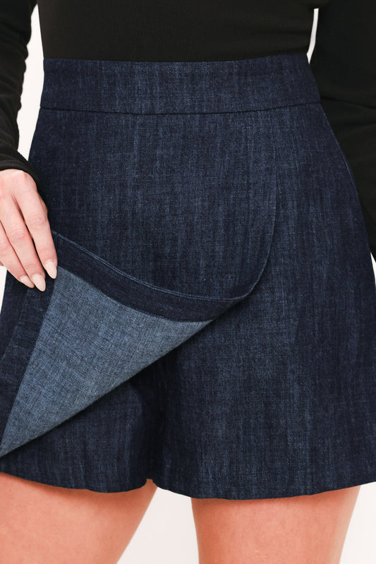 Close-up of a model wearing dark blue denim shorts with a matching denim lining shown being pulled up, paired with a black long-sleeve top.