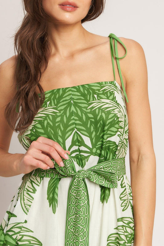 White and green woven fabric jumpsuit with a palm leaf print, tie shoulder straps, and a patterned waist bow.