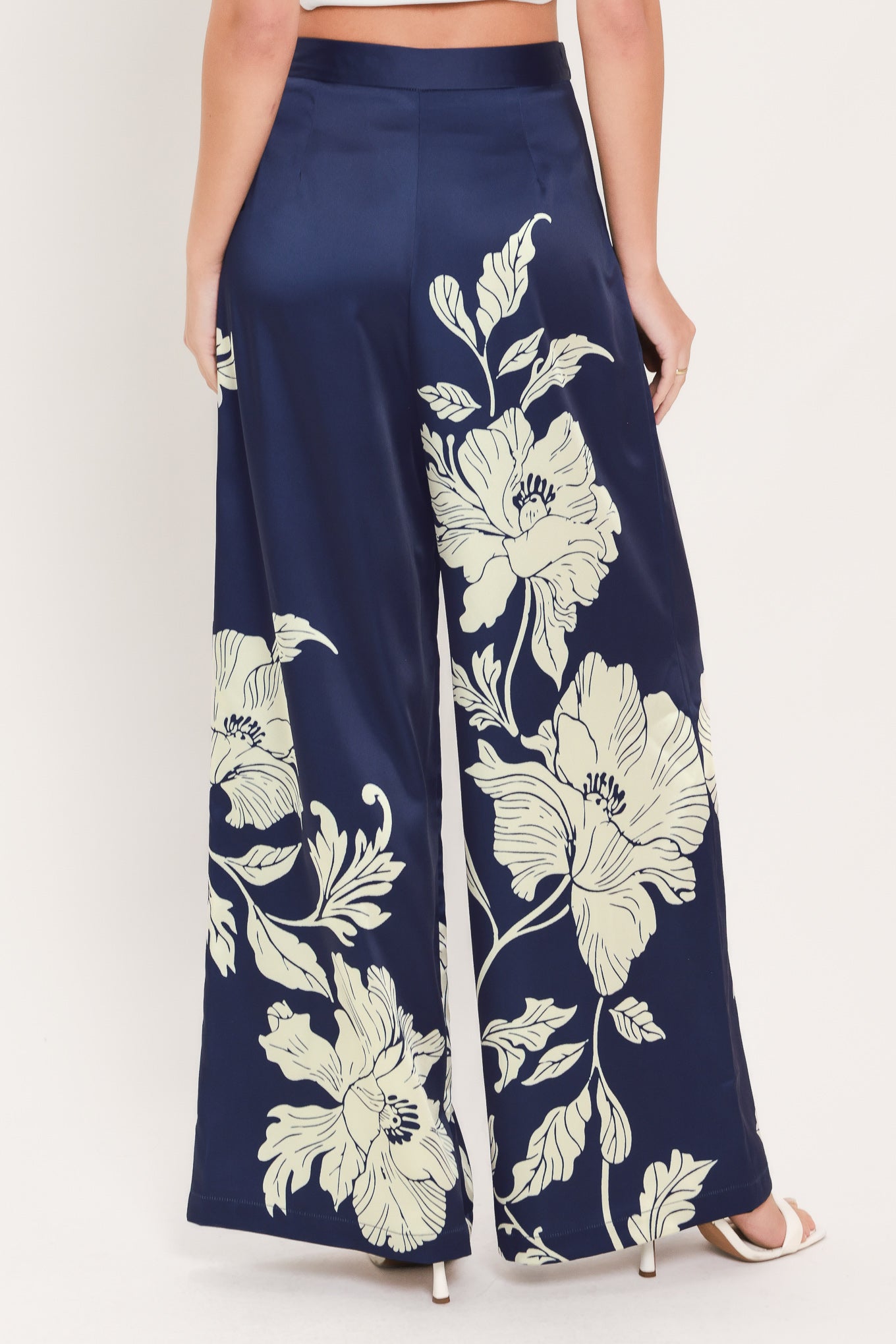 A model is shown from the waist down wearing navy blue wide-leg pants with a large cream floral print, paired with white heeled sandals against a light beige studio background.