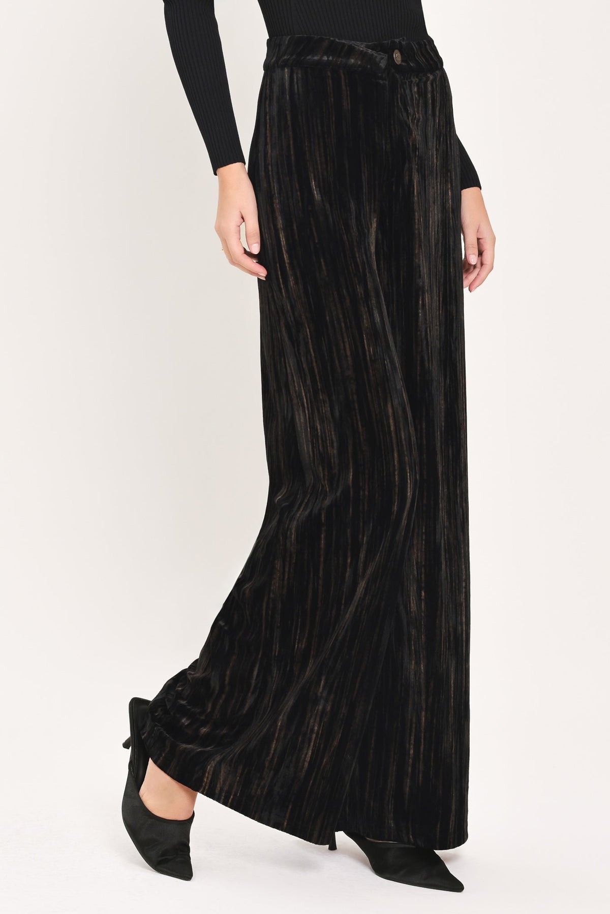 A model wearing black pleated velvet wide-leg trousers with a matching waist tie, styled with a black long-sleeve top and black pointed-toe heels against a plain white studio background.