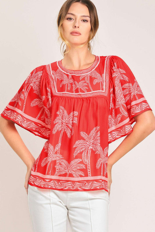Red short-sleeved fabric top with white palm tree print and geometric borders worn with white pants.