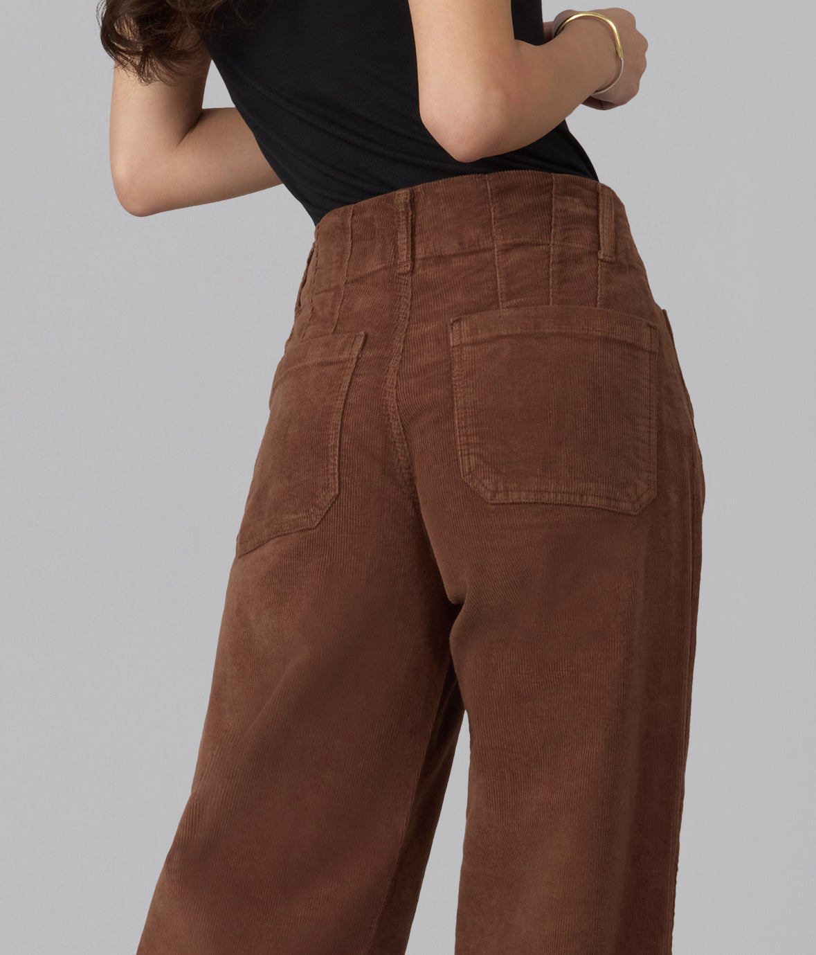 A close-up back view of a person wearing high-waisted brown corduroy pants with a black top, set against a solid gray studio background.