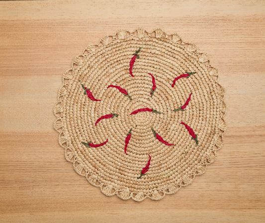 Scattered chili handwoven raffia round placemat with red chili pepper accents on a wooden surface.