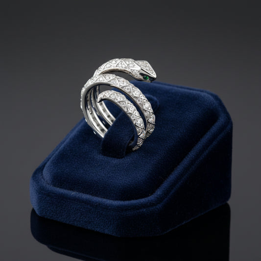 Coiled silver metal Silas Snake Ring with clear round stones and a green stone eye on a blue velvet ring display.
