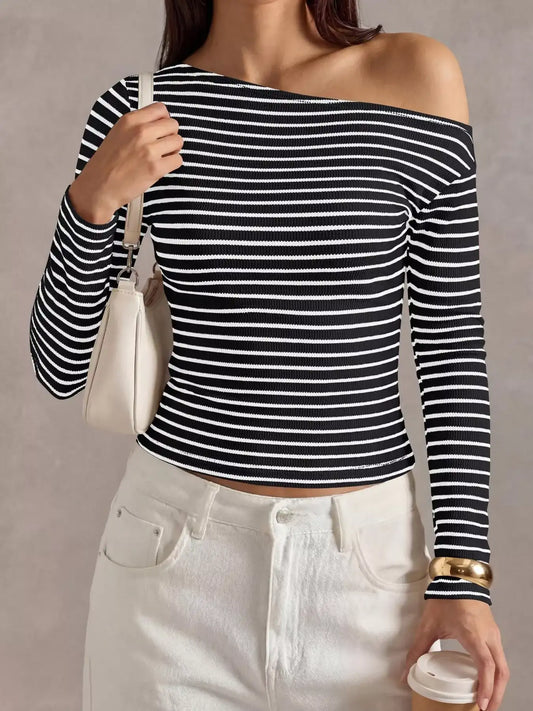 A woman wearing a black and white horizontally striped off-the-shoulder long-sleeve crop top, paired with white jeans, holding a white shoulder bag and a coffee cup against a neutral gray background.
