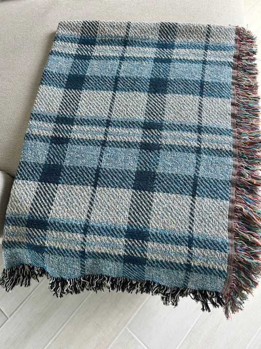 A woven plaid throw blanket in shades of blue, gray, and white, featuring multicolored fringe on one side, laid out on a light-colored sofa over a light wood floor.