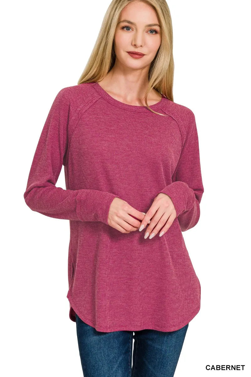 A woman modeling a heathered magenta long-sleeve top with raglan sleeves and a relaxed fit, paired with blue jeans against a plain white background. The text 'CABERNET' is visible in the bottom right corner.
