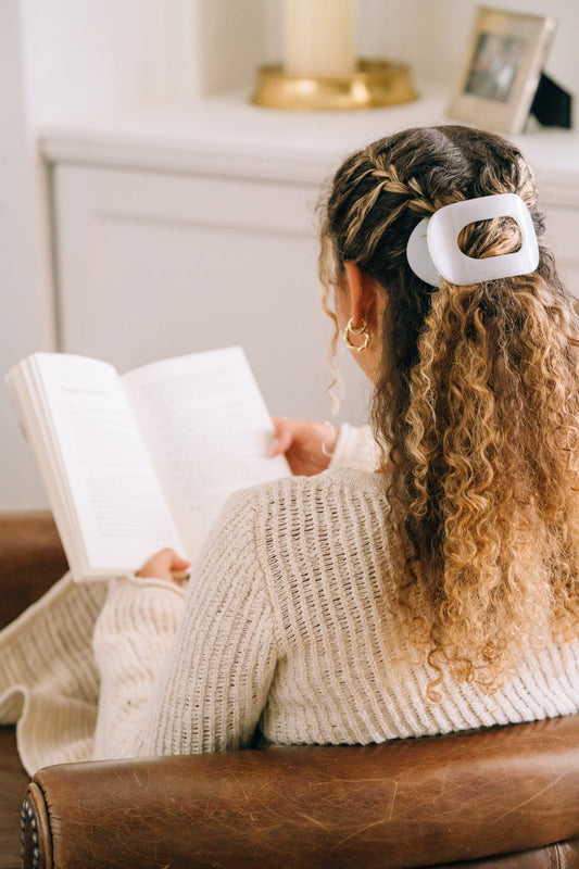 One white flat round plastic hair clip holding back curly hair on a person reading a book.