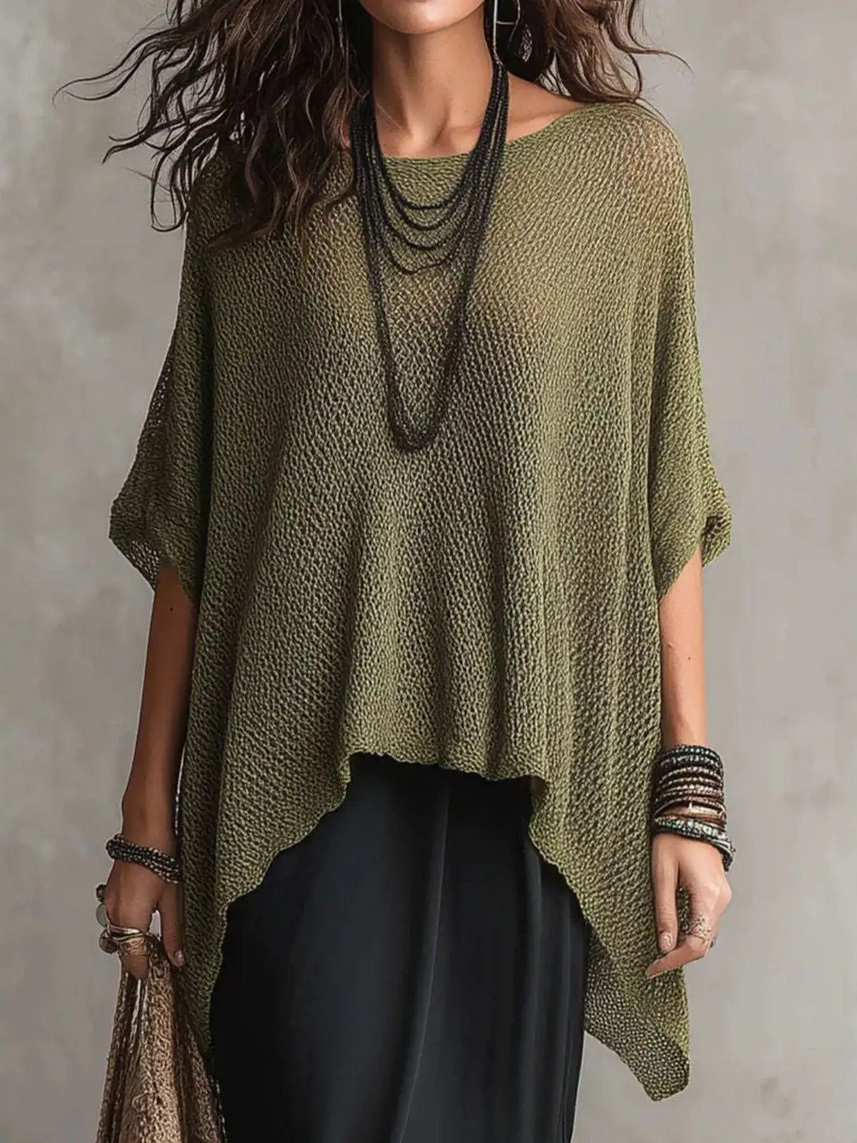 A woman wearing a loose, olive green open-knit poncho-style top with short wide sleeves, layered with long dark necklaces and bracelets, paired with black bottoms against a textured gray wall.
