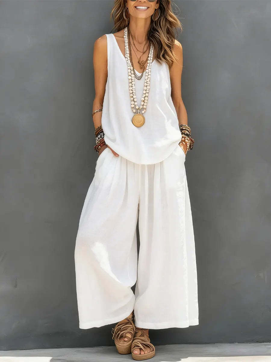 Woman wearing a white sleeveless top and wide-leg pants with layered necklaces against a gray background